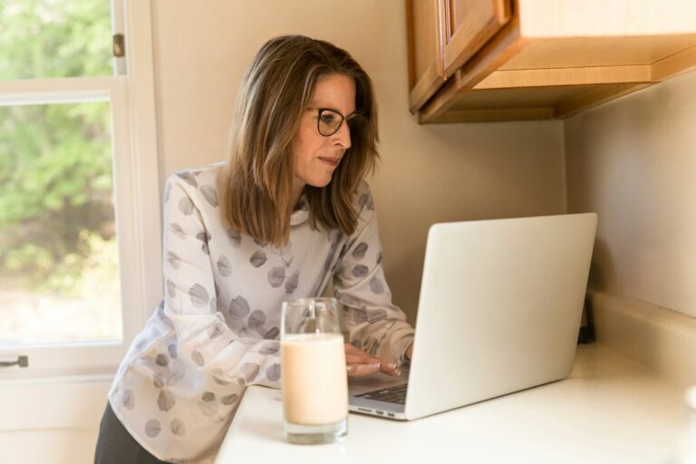 Woman with glasses using a laptop at a kitchen counter.