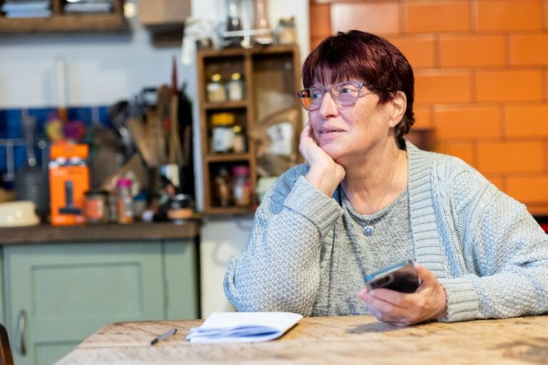 A woman holding a smartphone beside a notebook at a kitchen table.