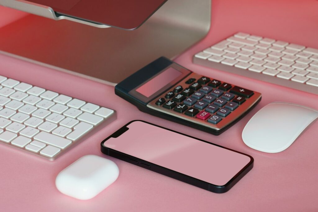 A calculator and blank smartphone screen beside computer accessories on a pink desk.