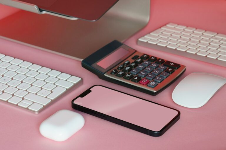 A calculator and blank smartphone screen beside computer accessories on a pink desk.