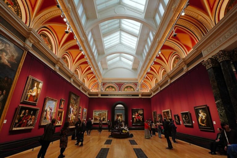 Visitors looking at framed paintings in an ornate museum gallery with red walls.