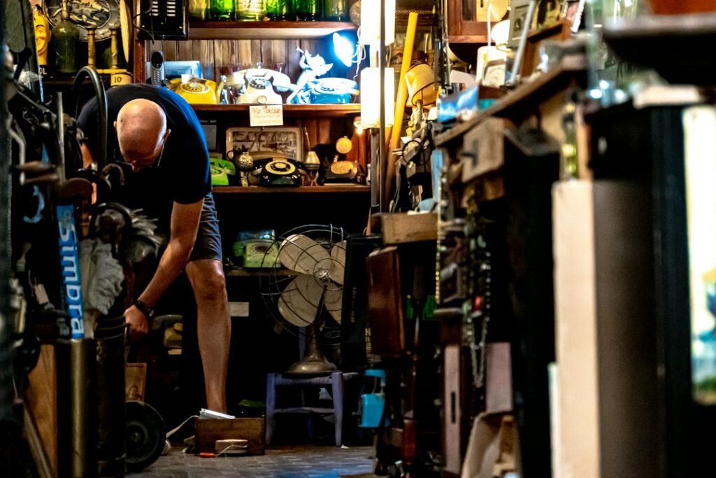 A man bending beside shelves of telephones, fans, and other items in a shop