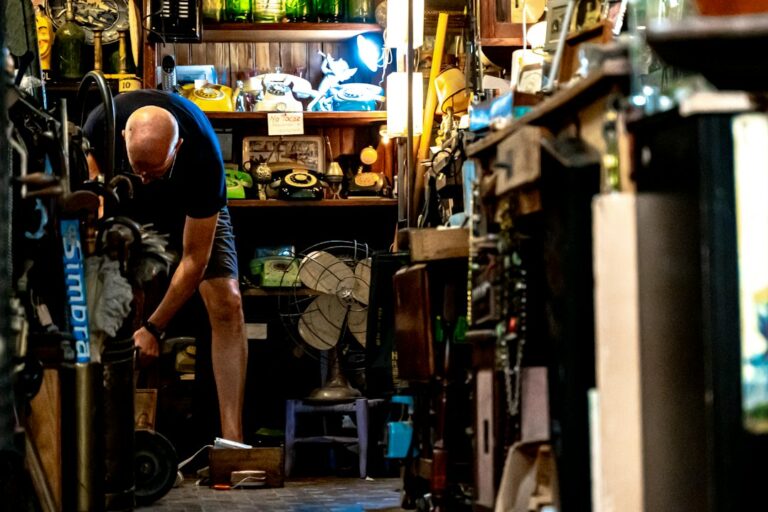 A man bending beside shelves of telephones, fans, and other items in a shop