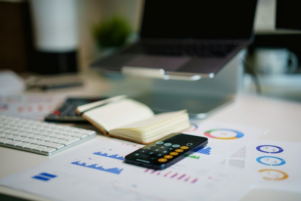 A smartphone calculator on printed charts beside an open notebook, keyboard and laptop.