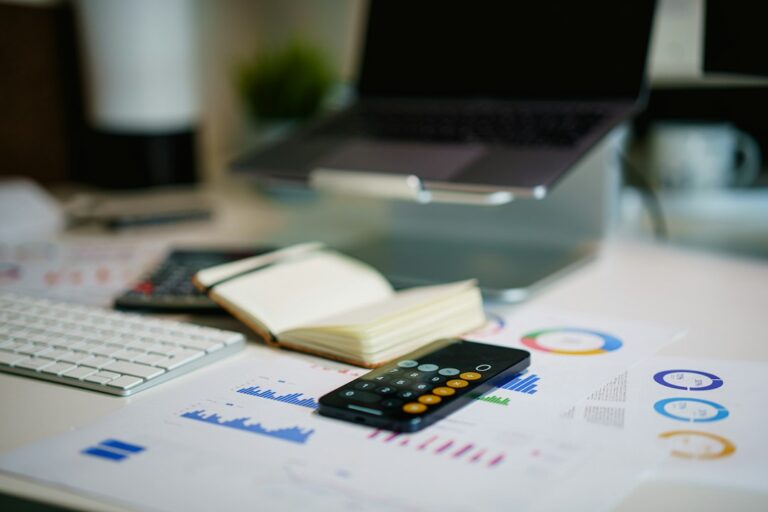 A smartphone calculator on printed charts beside an open notebook, keyboard and laptop.