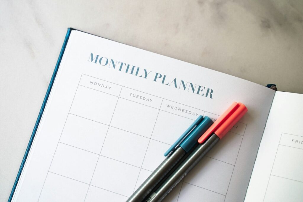An open monthly planner with teal and coral pens resting across its blank calendar grid.