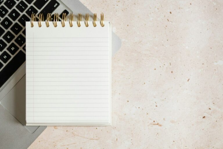 A blank lined notebook resting on part of a laptop on a pale desk.