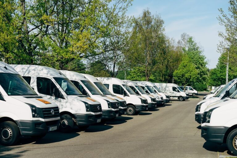 Rows of white vans parked in a paved lot with trees behind them.
