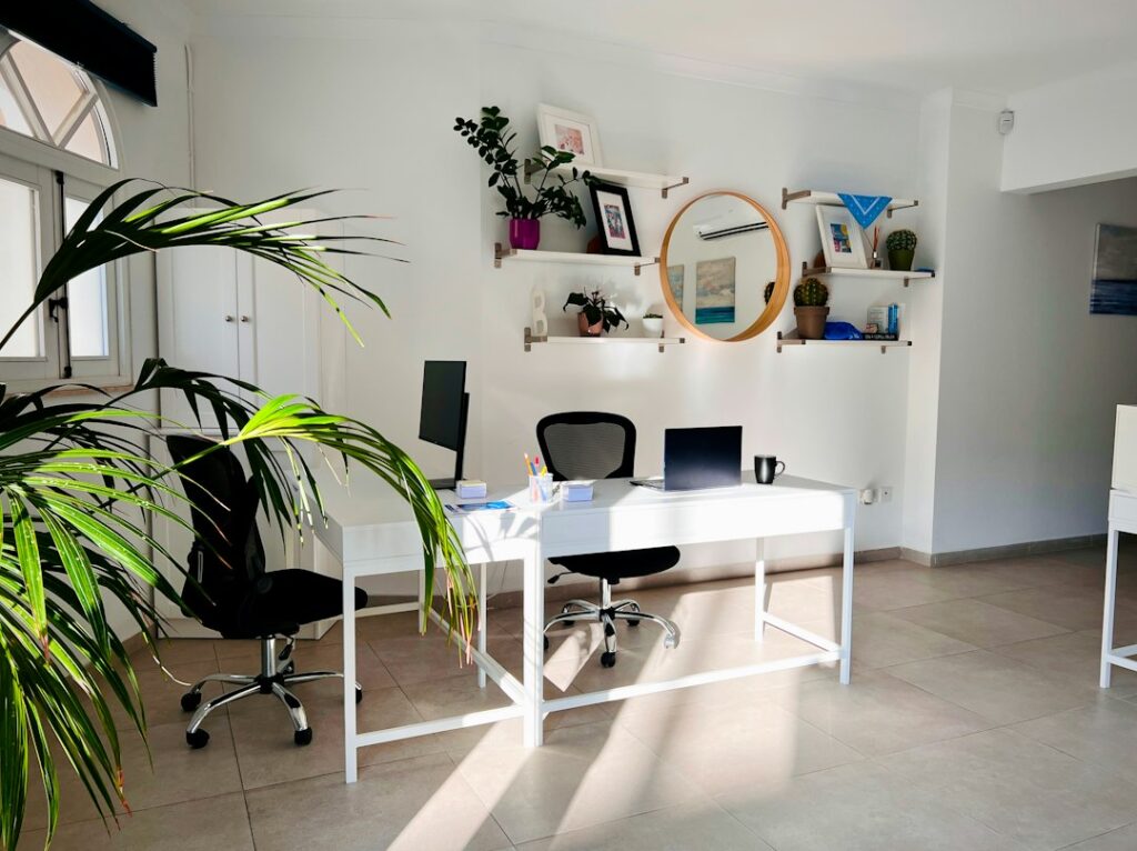 A bright office with a white desk, laptop, monitor, plants, and black chairs.