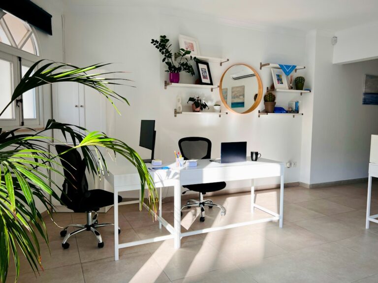 A bright office with a white desk, laptop, monitor, plants, and black chairs.