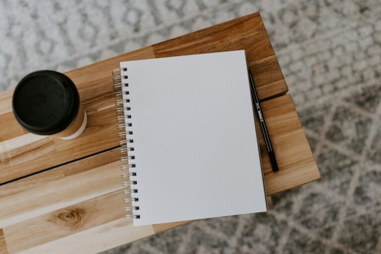 An open blank notebook, pencil and takeaway cup on a wooden bench.