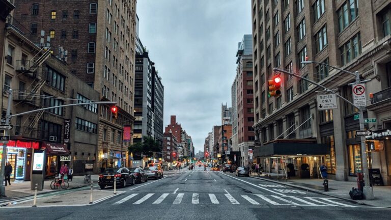 A city street lined with multistory buildings, storefronts, parked cars, and traffic lights.