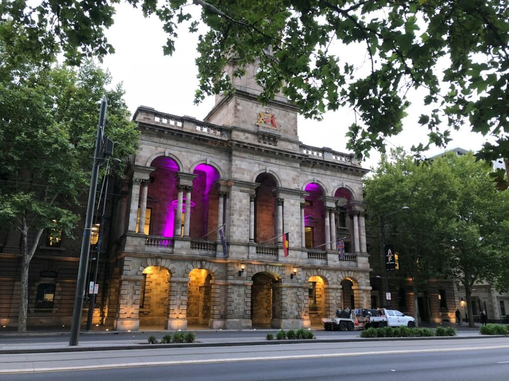 Stone civic building with arched windows and purple lighting beside a tree-lined street.