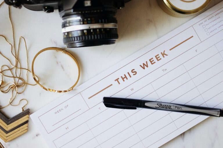 A weekly paper planner with a black pen, camera, and jewelry on a light surface.