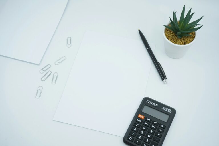 Blank paper with a calculator, black pen, paper clips, and a small potted plant.