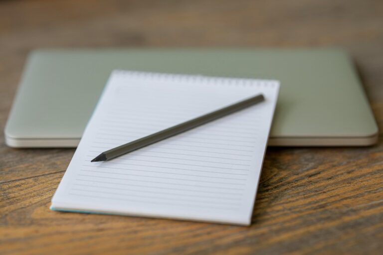 A pencil resting on a lined notebook in front of a closed laptop on a wooden table.