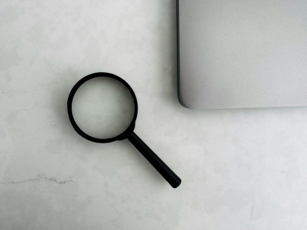 A black magnifying glass beside the corner of a silver laptop on a light surface.