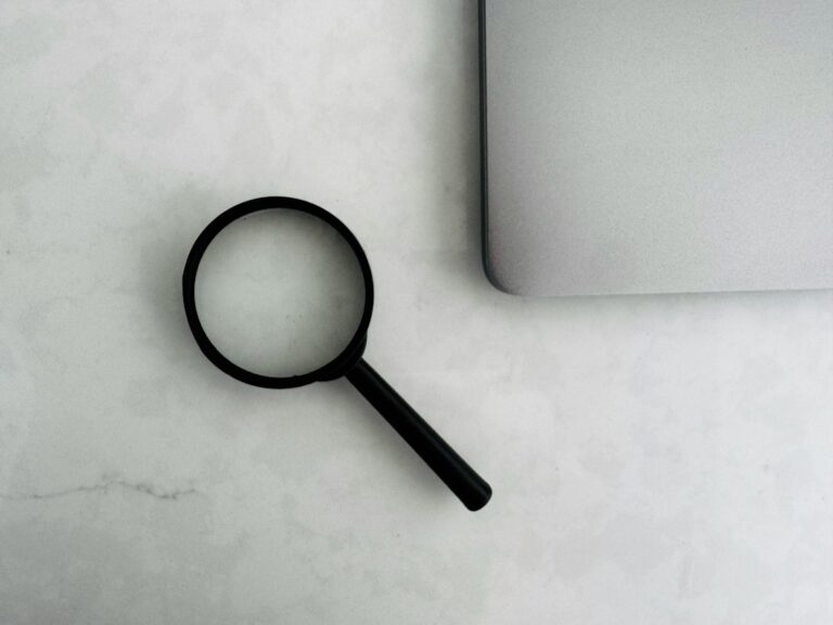 A black magnifying glass beside the corner of a silver laptop on a light surface.