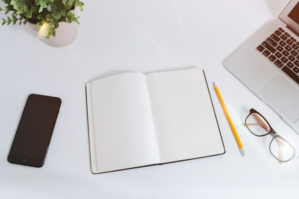 An open notebook, pencil, glasses, phone and laptop on a white desk.