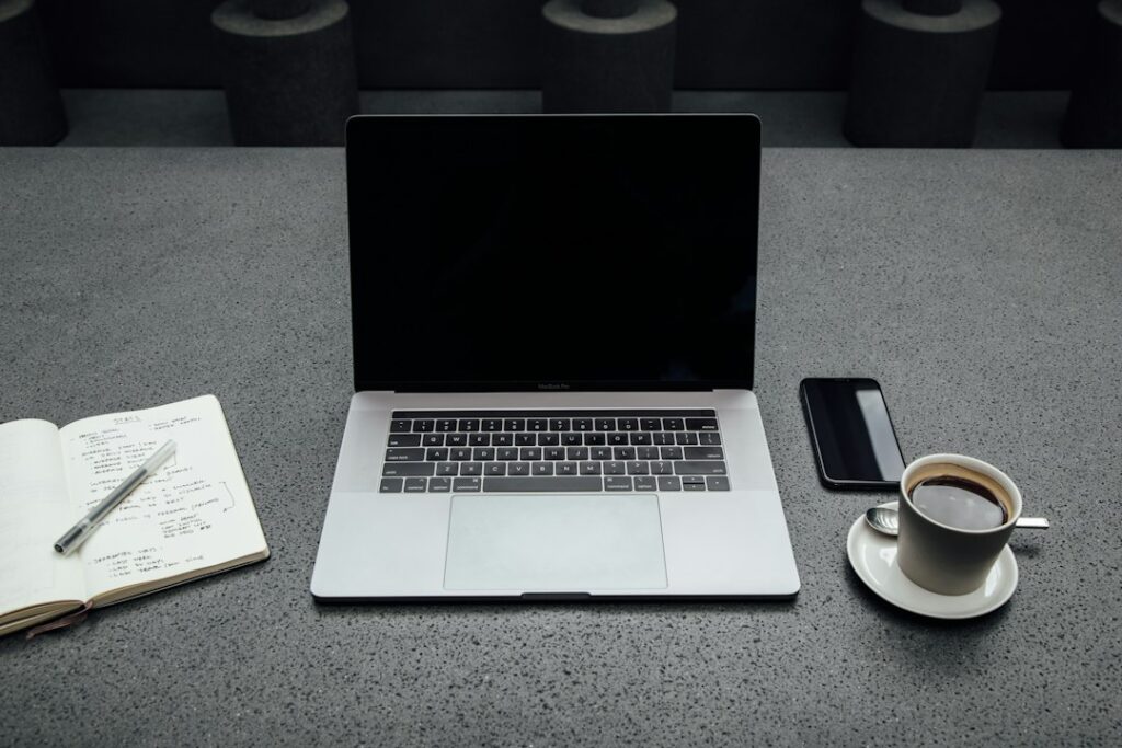 Laptop with a dark screen beside a notebook, phone and cup of coffee.