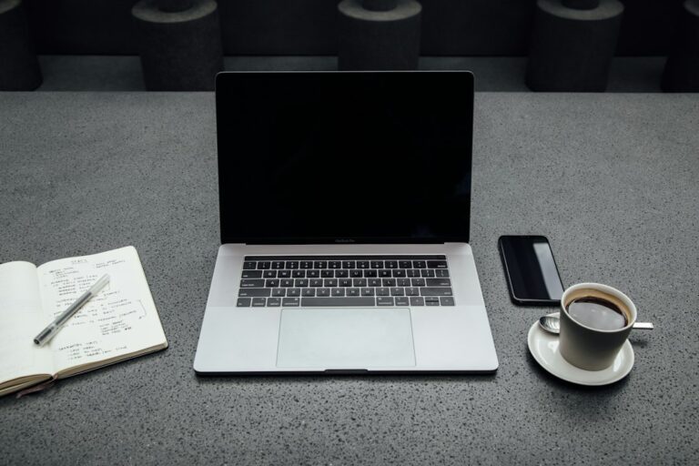 Laptop with a dark screen beside a notebook, phone and cup of coffee.