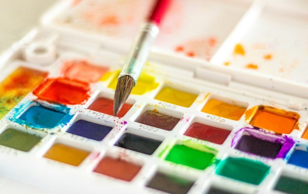 A paintbrush resting across a tray of colorful watercolor paints.