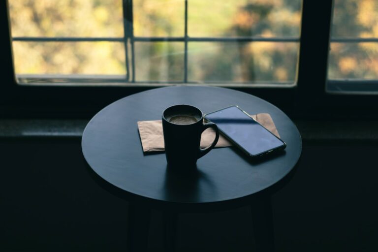 A mug and a smartphone on a small round table beside a window.
