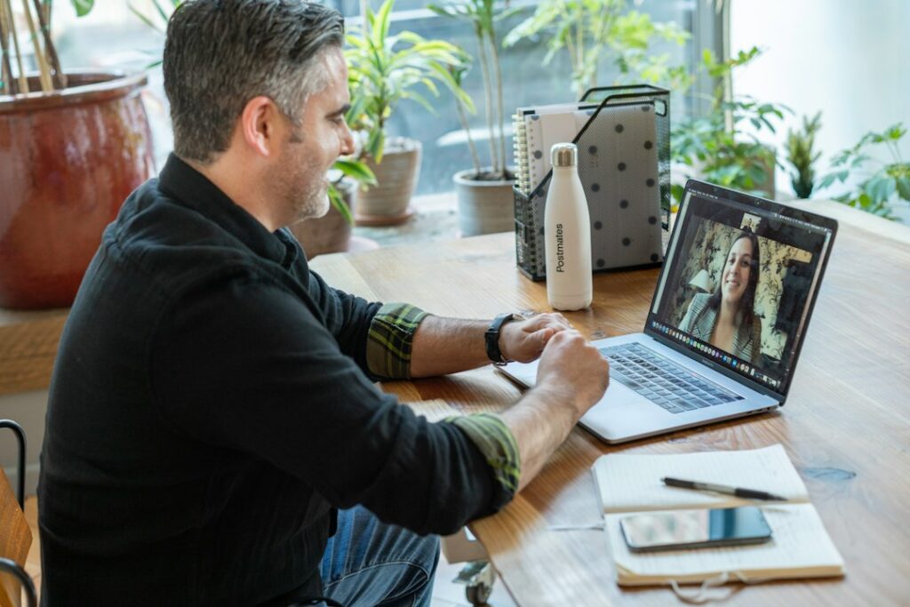 A man seated at a desk speaking with a woman on a laptop video call.