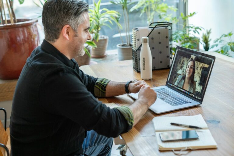 A man seated at a desk speaking with a woman on a laptop video call.