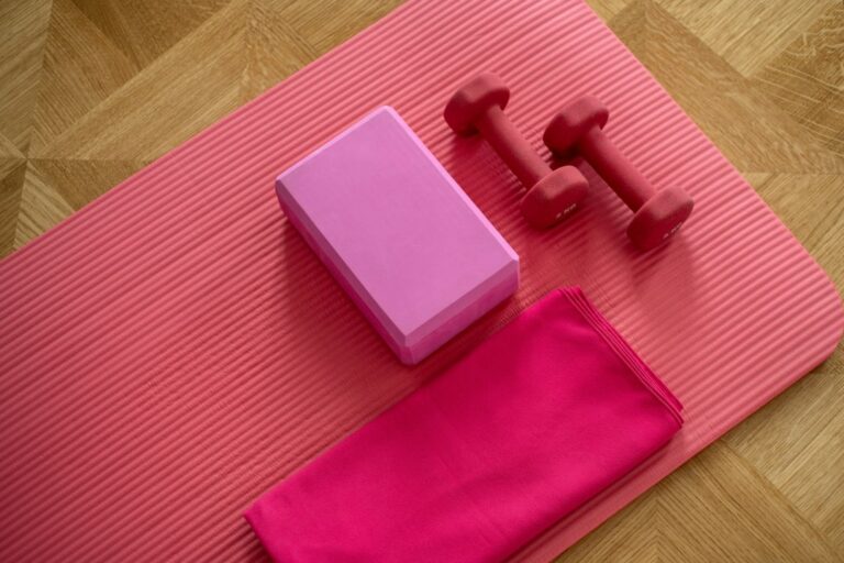 Two red dumbbells, a yoga block, and a folded towel on a pink exercise mat.