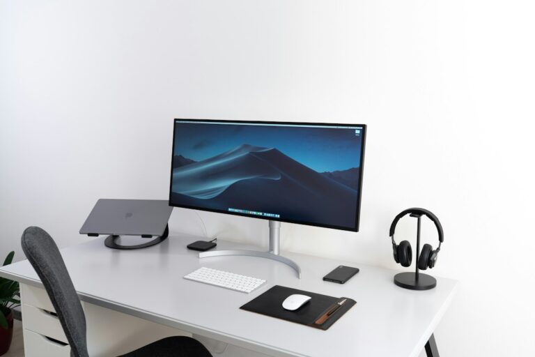 Desk with a monitor, keyboard, mouse, closed laptop and headphones beside an office chair.