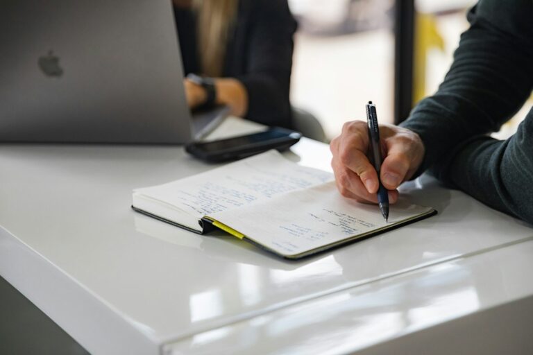A hand writing in an open notebook beside a laptop.