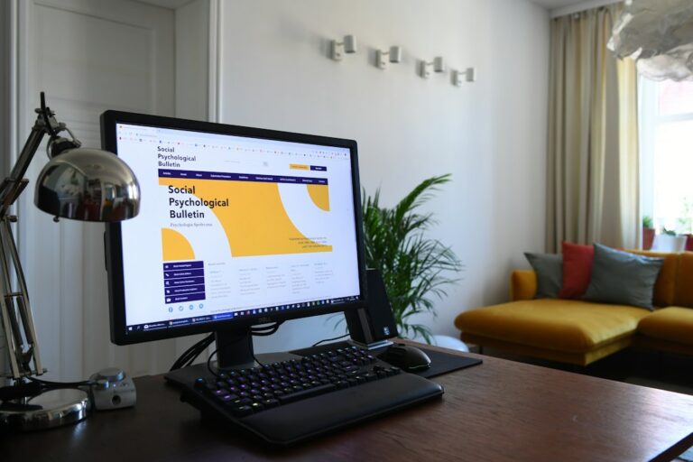 Desktop monitor, keyboard, and lamp on a desk beside a window.