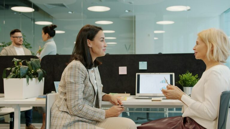 Two women talking beside a laptop in an office, with two colleagues in the background.