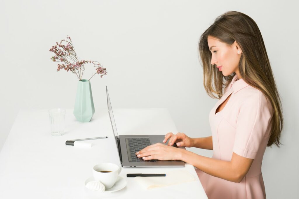 A woman using a laptop at a desk beside a notebook and cup.
