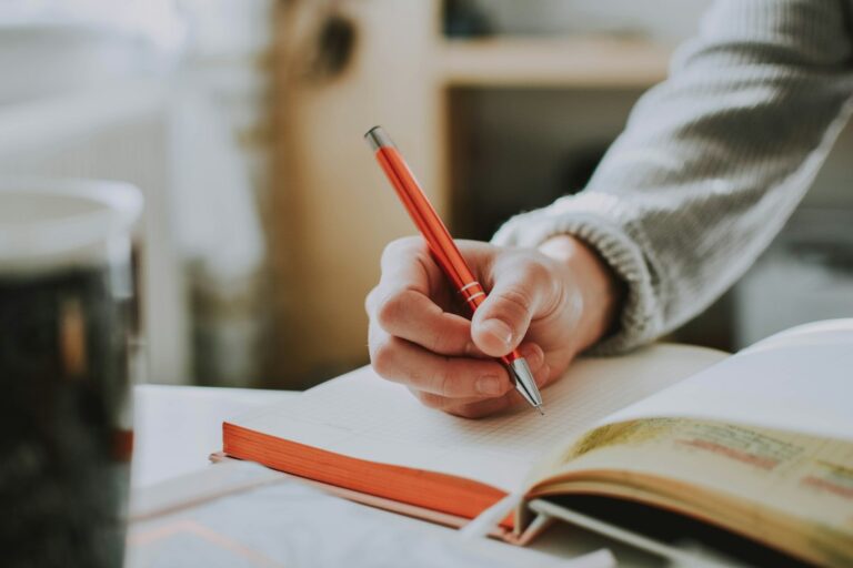 Close-up of a hand writing with a red pen in an open notebook.