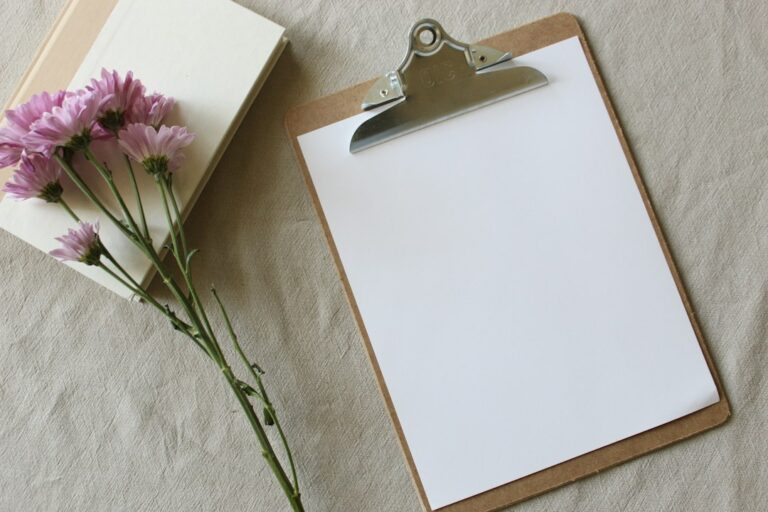 Blank paper on a clipboard beside a closed book and pink flowers.