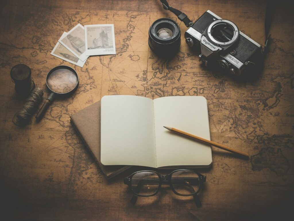 An open notebook and pencil on a map beside a camera and glasses.