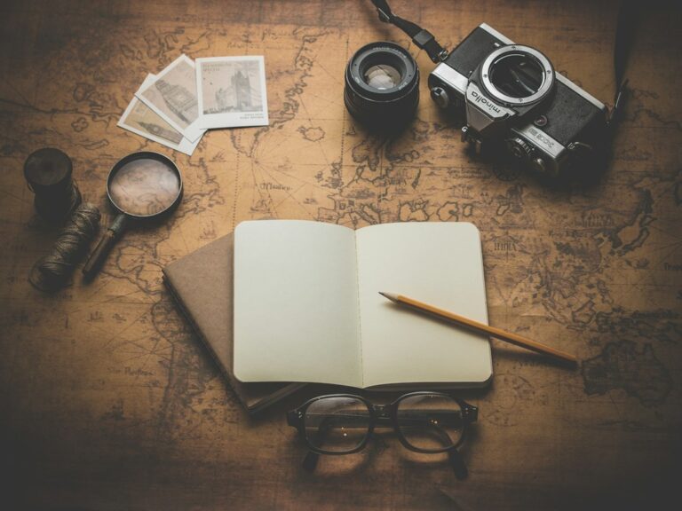 An open notebook and pencil on a map beside a camera and glasses.