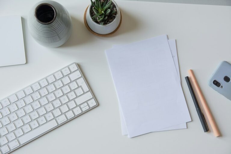 Blank sheets of paper with a keyboard, pens, phone and small plant on a desk.