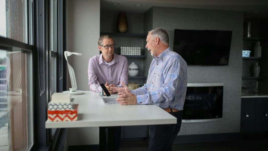 Two men discussing a laptop at a standing desk beside a window.