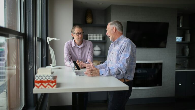 Two men discussing a laptop at a standing desk beside a window.