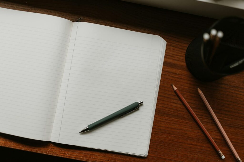 Open lined notebook with a green pen and two pencils on a wooden desk.