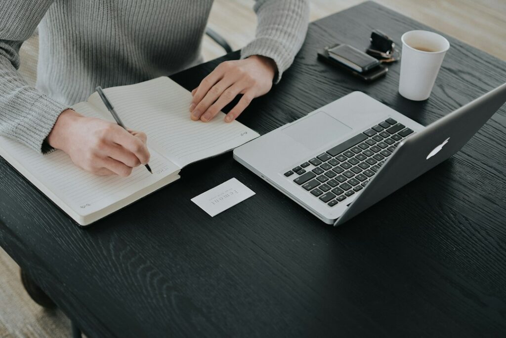 Close-up of hands writing in a notebook beside an open laptop.