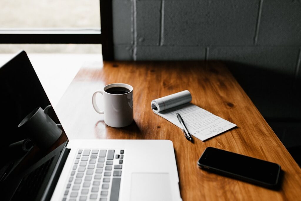 A laptop, coffee mug, notepad and phone on a wooden desk.