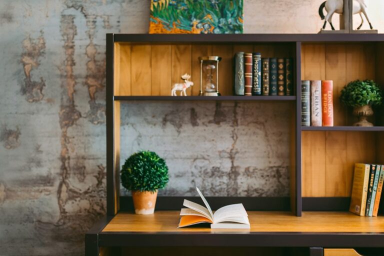 An open book on a wooden desk beneath shelves of books and plants.