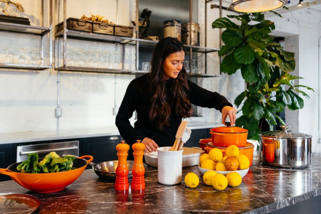 A woman checking a pot in a kitchen with vegetables, lemons and cookware.