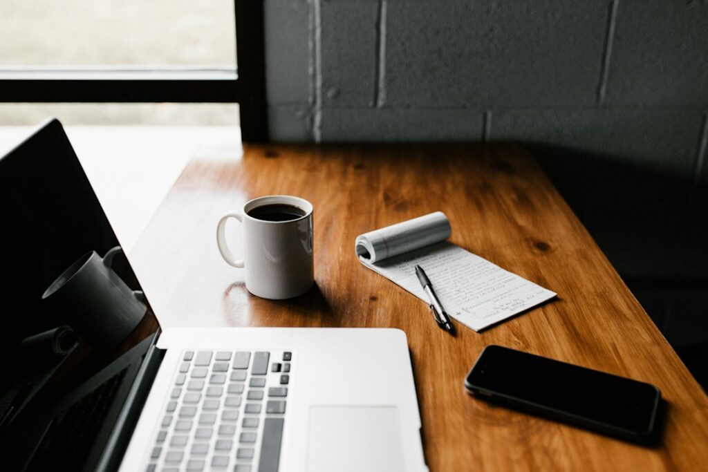 A laptop, handwritten notepad, coffee mug, and phone on a wooden desk.