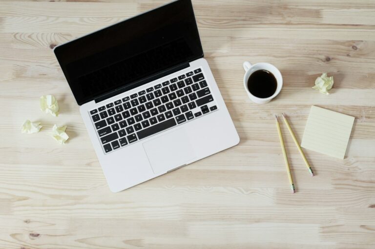 A laptop, cup of coffee, pencils, and a blank note on a wooden desk.