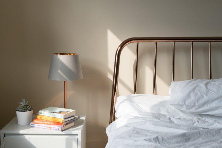 A bedside lamp, books, and white bedding in a sunlit bedroom